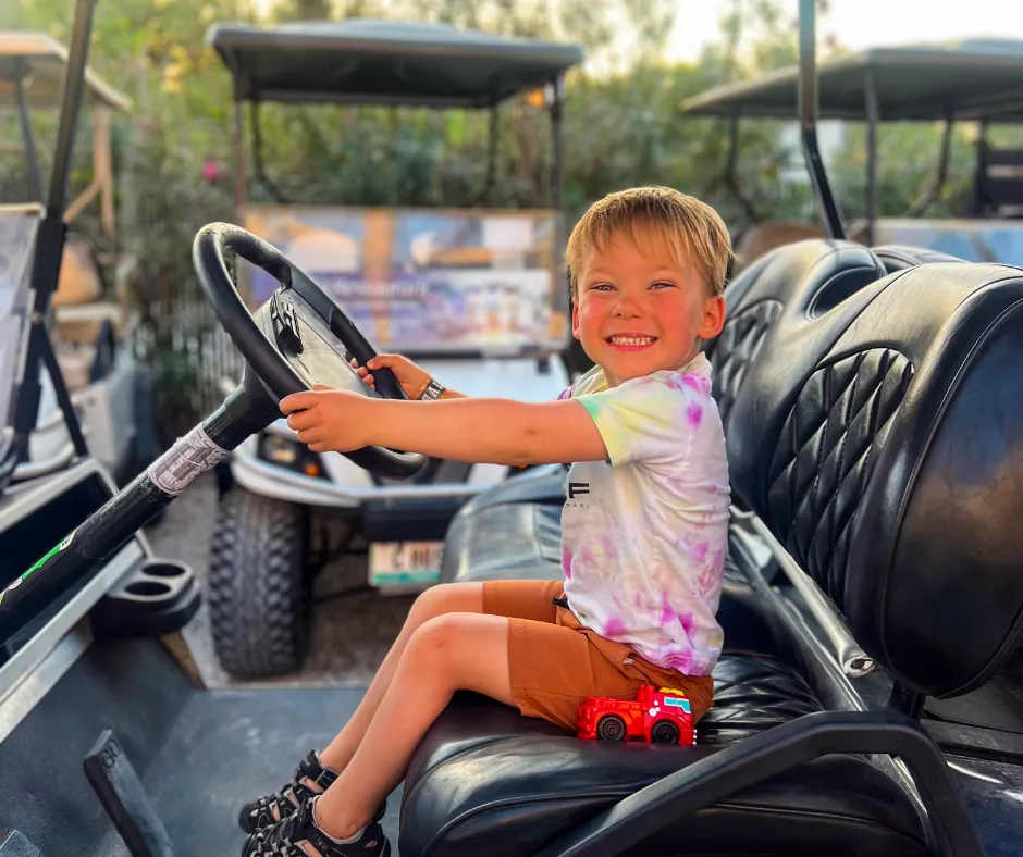 Joyful boy driving a golf cart at Belize Beach & Jungle Stays We Love With Kids, tie-dye shirt, red toy truck on lap.