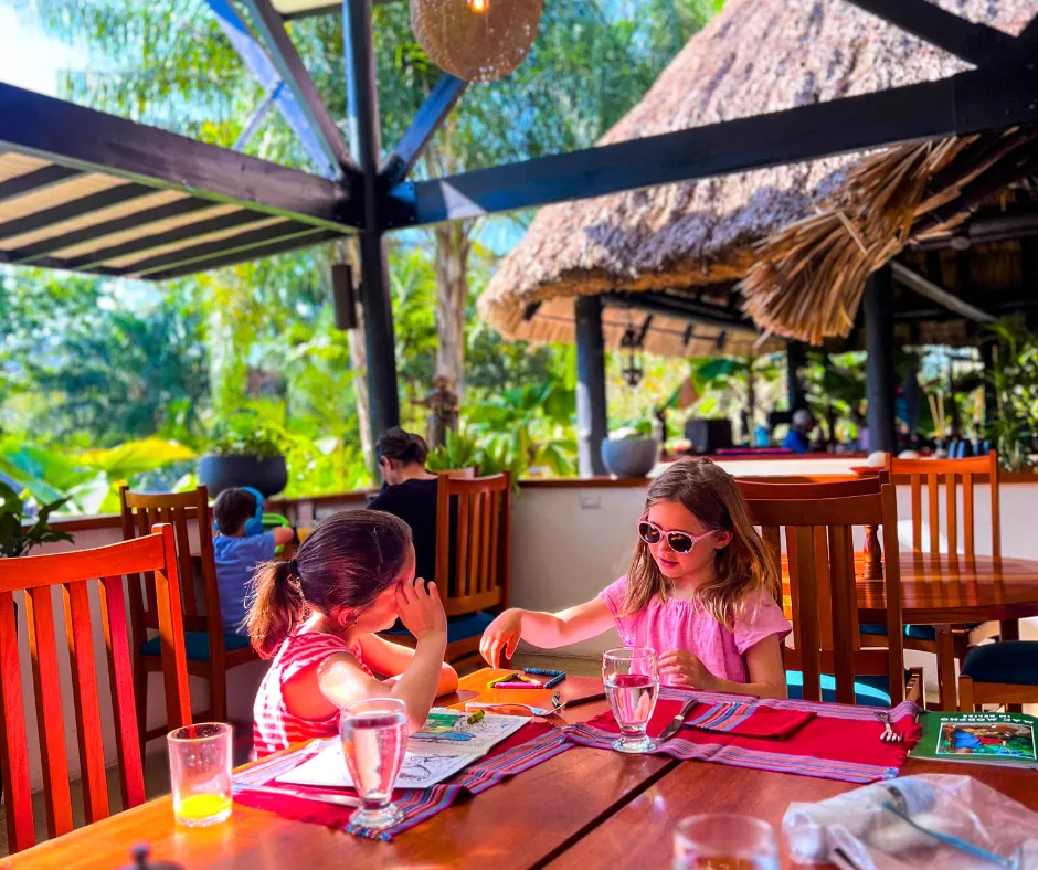Breakfast with kids at a Belize beach-jungle resort, two girls color at a wooden table under a thatched roof.