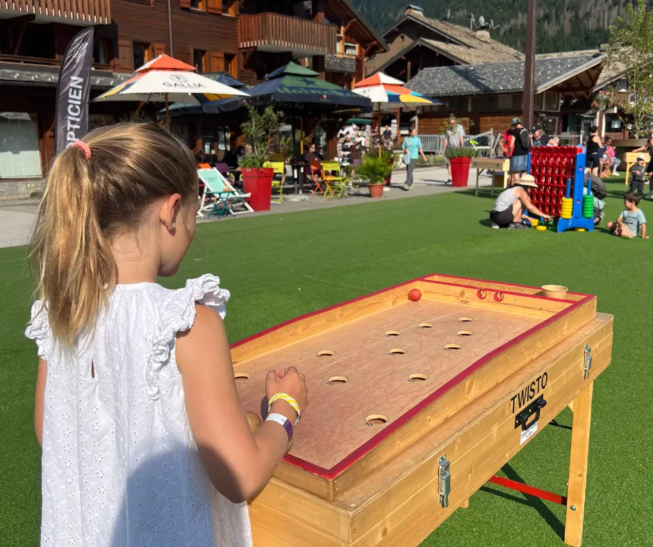 Young girl in a white dress plays a wooden tabletop game on artificial turf in a French Alps town square with chalet-style buildings.