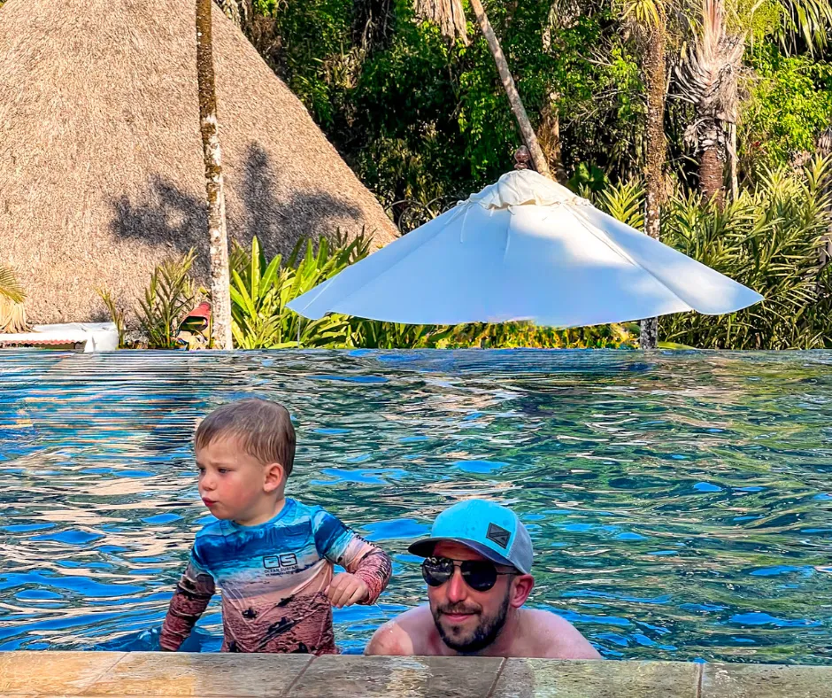 father and toddler in a tropical lodge pool, with a thatched hut and lush plants.