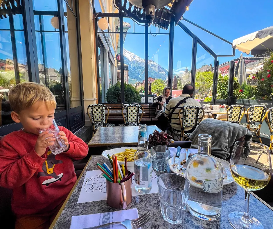 French Alps outdoor cafe scene with a child enjoying a kid-friendly lunch, sipping water near drawing supplies in Chamonix.