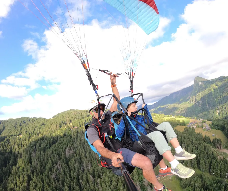 Tandem paragliding over the French Alps: child passenger and instructor in harnesses, soaring above lush forests.