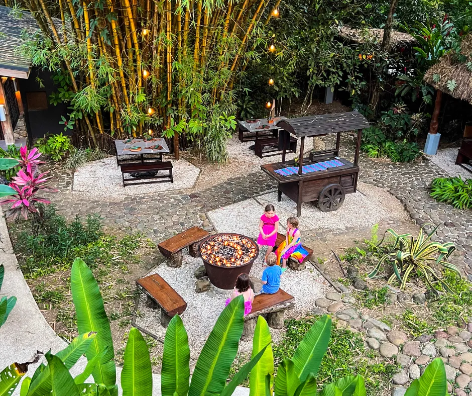 tropical lodge courtyard with a fire pit among bamboo, where kids sit on benches.