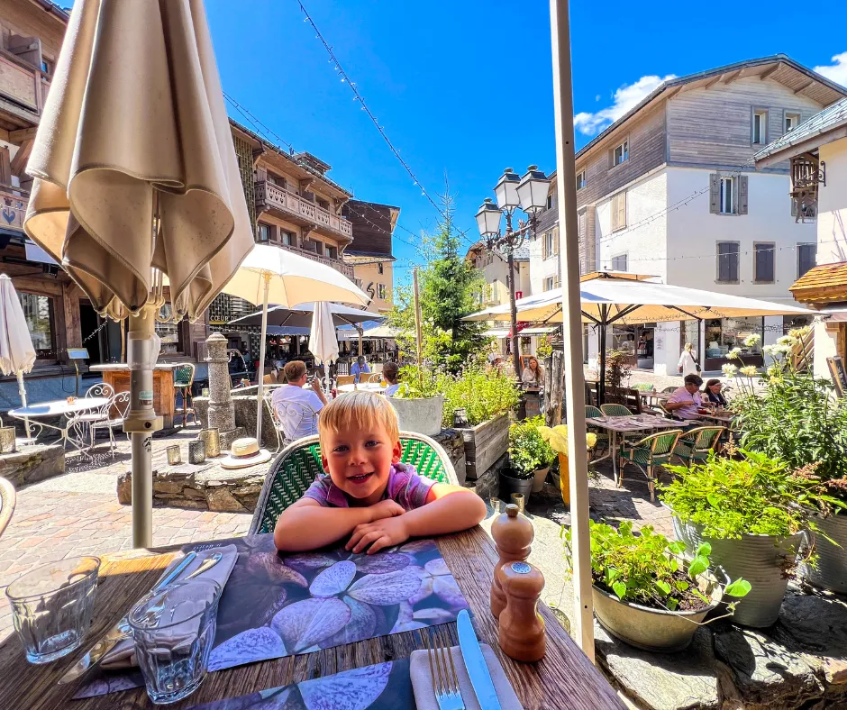 Young child at a sunny outdoor cafe lunch in Megeve, French Alps, with umbrellas, cobblestone square, and diners amid potted plants.