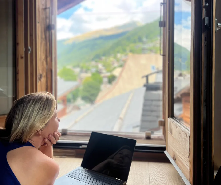 Mother planning a trip on a laptop, gazing from a wooden-window nook at French Alps village and mountains.