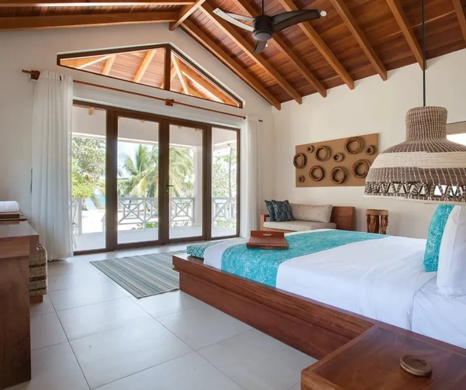 Belize beach resort bedroom with wood-beam ceiling, glass doors to a tropical balcony and turquoise bed runner.