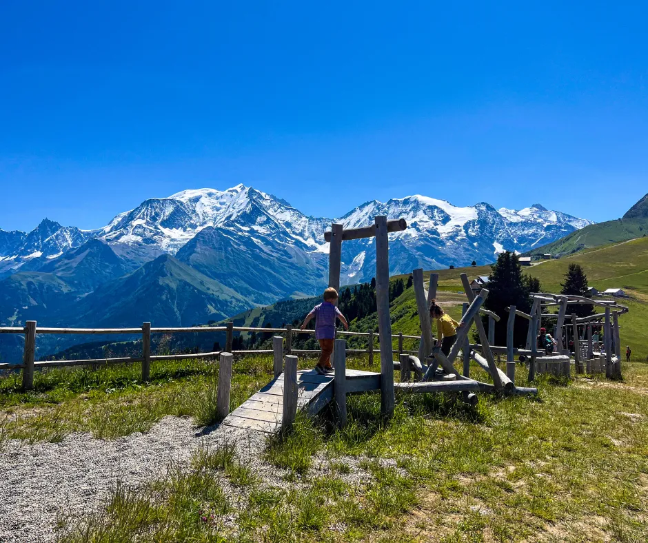 Children explore a rustic wooden playground on a grassy hillside, with snowcapped French Alps beneath a clear blue sky.